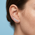 A close-up of a model's ear wearing a curved cluster of silver ear cuffs adorned with small clear stones, set against a solid light blue background.