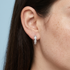 A close-up of a model's ear wearing a slim silver huggie hoop earring set with clear pavé gemstones, against a light blue background.
