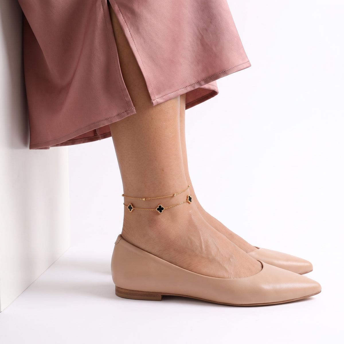 Side profile of foot in nude flats beside a white block wearing Lise gold clover anklet