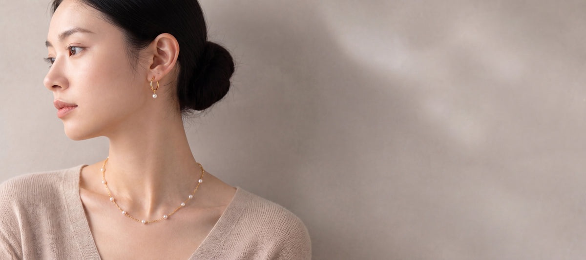 Woman in an oatmeal knit wearing a gold pearl necklace and small gold hoops